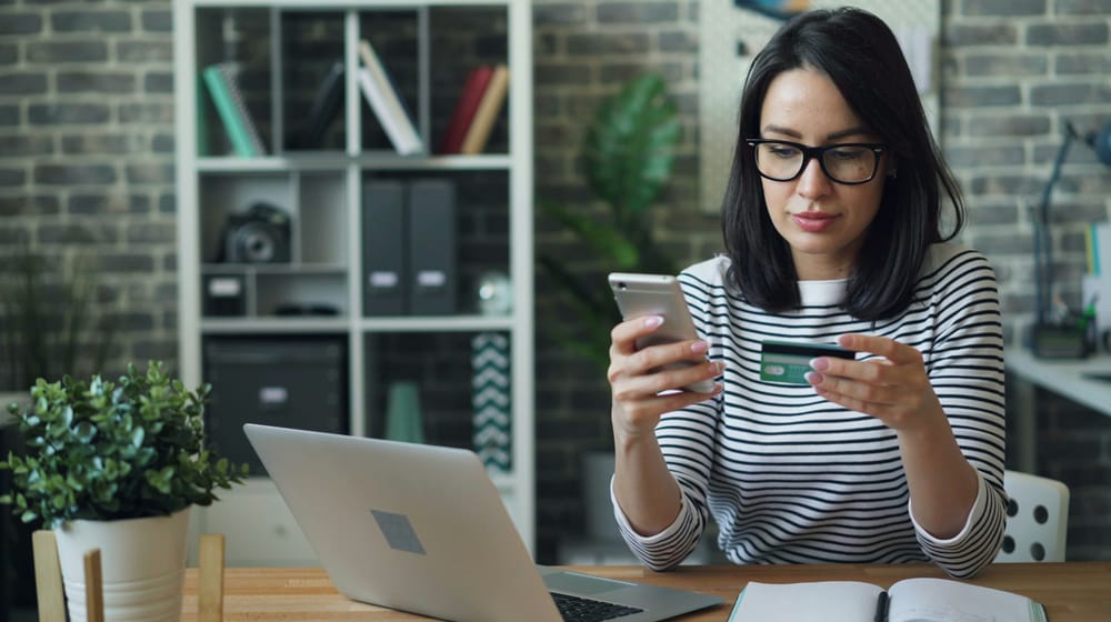 Woman at home shopping on her phone with a card in hand, laptop nearby